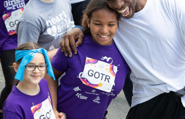Photograph of a GOTR participant smiling with her father. He hugs her and smiles. Participcant is wearing a purple GOTR program shirt and race bib that reads "GOTR". Father wears white shirt and black shorts.